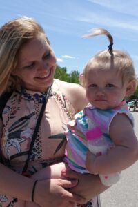 A woman holding a baby outdoors on a sunny day. The woman is smiling and looking at the baby, who is wearing a colorful striped dress and has a small ponytail on top of their head, while holding a bottle. They are in a bright, open area with a clear blue sky.