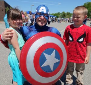 A person dressed as Captain America smiles and kneels, posing with two children. The child on the left, in a turquoise dress, smiles while the child on the right, in a Spider-Man T-shirt, also smiles. They are outdoors in a parking lot with other people in the background.