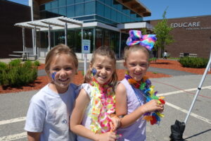 Three young girls smile brightly while standing outside a modern building with large glass windows. They are wearing colorful leis and one has a large, vibrant bow in her hair. The middle girl has a painted butterfly on her cheek.