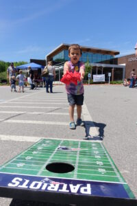 A young boy, wearing a tie-dye shirt and blue shorts, throws a bean bag at a cornhole board with "PATRIOTS" written on it in a parking lot. People are seen in the background near buildings and tents on a sunny day.