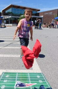 A young child wearing a tie-dye t-shirt and shorts throws a red fabric beanbag towards a target on the ground in an outdoor setting. The target has a football field design with a New England Patriots logo. A modern building labeled "EDUCARE" is in the background.