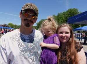 A man with a brown beard wearing a white graphic t-shirt and a cap, stands next to a woman with long, light brown hair. Both are smiling. The woman holds a young girl in a purple dress who is looking down, also smiling. They are outdoors on a sunny day.