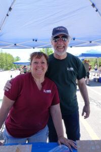 Two smiling people stand under a canopy at an outdoor event. The woman, wearing a red T-shirt, stands on the left while the man, in a green shirt and baseball cap, is on her right with his arm around her shoulder. Other canopies and people are visible in the background.