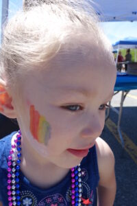 A child with light hair in a bun, wearing a blue top and colorful beaded necklaces, has a painted rainbow on their cheek. The child is standing outdoors under a tent on a sunny day.