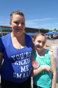 A woman and a young girl stand side by side outdoors on a sunny day, both smiling at the camera. The woman wears a blue shirt with the text "YOU'RE KICKING ME SMALLS." The girl wears a green tank top and holds a drink with a red straw.