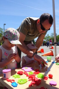 Two children wearing sun hats are engrossed in crafting with colorful playdough at a table outside on a sunny day. An adult, wearing sunglasses and a brown t-shirt, assists them by arranging the playdough shapes. A traffic cone and people are visible in the background.