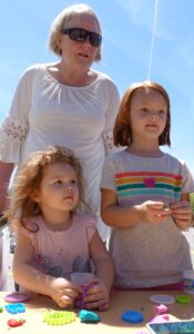 An older woman with gray hair and sunglasses stands behind two young girls with red hair. The girls are engaged in playing with colorful modeling clay at an outdoor table. The sky is clear and blue in the background.