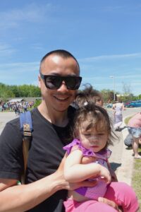 A man wearing sunglasses and a black shirt holds a young girl with sunglasses on her head in his arms. They are standing outdoors under a clear blue sky, with other people and trees visible in the background. The child is dressed in a pink outfit.