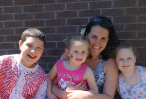 A woman and three children smile at the camera in front of a brick wall. The boy on the left wears a white shirt with red patterns, the girl in the middle has face paint and a pink shirt, and the girl on the right has curly hair and a blue shirt.
