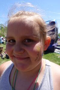 A young child with face paint smiles at an outdoor event. The face paint features a sun and squiggly lines. The child is wearing a gray top and colorful necklace. In the background, people and an inflatable structure are visible under a clear blue sky.