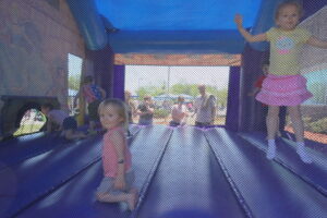 Children are playing inside a blue and purple inflatable bounce house. One child is kneeling and looking at the camera, while another is jumping with arms raised. Several adults and other children are visible in the background outside the entrance.