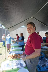 A man wearing a red shirt and gloves is smiling while serving food under a large tent. He is standing in front of trays filled with burgers. Other people are also serving or getting food in the background. It appears to be a community or outdoor event.