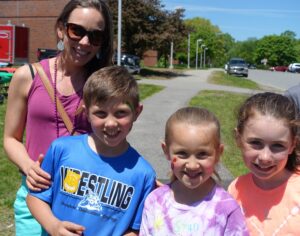 A woman and three children smile outdoors on a sunny day. The woman wears sunglasses, a pink necklace, and a light purple top. A boy in a blue "Wrestling" shirt stands next to two girls, one with a heart mark on her cheek, all wearing casual clothes. Trees and a pathway are in the background.