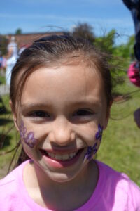 A smiling young girl with face paint stands outdoors. She has dark hair and is wearing a pink shirt. Purple flowers are painted on her cheeks. The background is a sunny day with green grass and people in the distance.