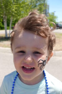 A young child with short, tousled hair smiles outdoors. They have a painted black spider on their cheek and are wearing blue bead necklaces. The background features greenery and a paved area.