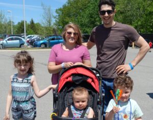 A family of five stands outdoors in a sunny parking lot. A man and woman smile with their arms around three children. One child stands, one is in a stroller, and one holds colorful balloon sticks. Cars and green trees are visible in the background.
