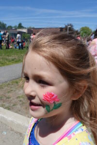 A young girl with blond hair is outdoors at a park or festival. She has a painted pink and green rose design on her cheek. The background shows other people and trees under a clear blue sky. She is smiling and looking to the side.
