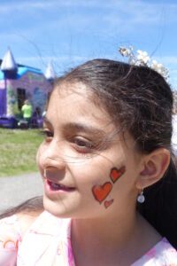 A young girl smiles outdoors with her cheek painted with three red hearts. She has long hair adorned with a floral headband, and she's wearing earrings. In the background, an inflatable structure and a person in a green shirt are visible against a bright blue sky.