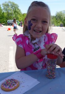 A young girl with a painted paw print on her cheek smiles while decorating a cookie with colorful sprinkles. She wears a pink shirt with white dots and a flower lei, sitting at a table outdoors on a sunny day. Trees and outdoor event activities can be seen behind her.
