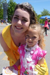 A woman in a yellow dress and tiara poses with a smiling young girl in a pink dress and white lei. The girl holds a sprinkled donut. They are outside at an event, with a climbing wall and other people visible in the background.