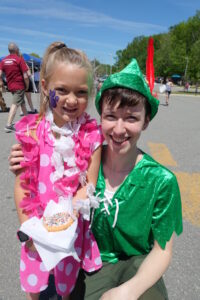 A young child in a pink polka dot dress and a floral lei stands beside an adult dressed as Peter Pan. The child has a purple butterfly painted on their cheek and is holding a sprinkled donut. It's a sunny day with people and trees in the background.