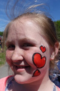 A girl with light brown hair smiles outdoors. Her face, which has freckles, features three painted red hearts of varying sizes on her right cheek. She’s wearing a light pink top, and a clear blue sky is in the background.