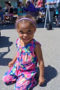 A young girl dressed in a colorful, tie-dye sundress smiles at the camera while crouching on a paved area. She has a headband in her hair and outdoor people and activities can be seen in the background.