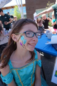 A young girl with glasses smiles at the camera, showing off face paint of colorful balloons on her cheek. She is wearing a blue dress with yellow trim. In the background, people are seated at a table under a canopy outside.