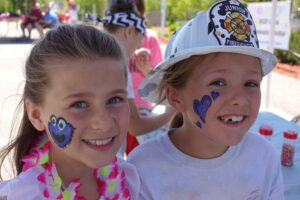 Two young girls with face paintings smile at the camera. The girl on the left has a blue monster face painted on her cheek, while the girl on the right has a purple heart. The girl on the right is also wearing a white junior firefighter helmet.