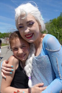 A smiling woman dressed as Elsa from Disney's "Frozen" hugs a young girl with braces and painted nails. They are outdoors on a sunny day, and the girl has a blue star painted on her cheek. Both appear happy and excited. Green trees and a blue sky are in the background.