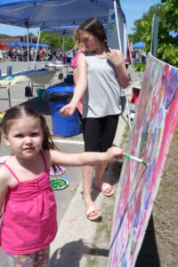 Two children engage in a painting activity outdoors at an event. One child in a pink top paints on a large colorful canvas, while an older child in a gray top and black pants watches. People, tents, and tables can be seen in the background.