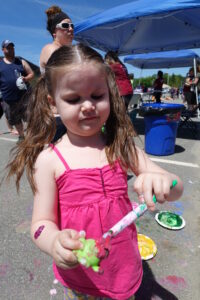 A young girl with pigtails, wearing a pink top, paints a small green toy with a brush at an outdoor event. Other people are gathered under a blue canopy in the background. Paint supplies are visible on the ground.