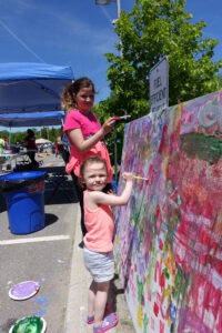 Two young girls, one in a pink shirt and the other in a light pink top, enthusiastically paint a large, colorful mural outdoors on a sunny day. They are standing on a sidewalk near a "Fuel Efficient Vehicle" sign with a blue tent canopy and green trees in the background.