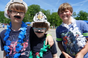 Three children are dressed up and standing together outdoors on a sunny day. Two of them wear hard hats, one with a "Junior Firefighter" badge, and fake mustaches. They all have leis around their necks and are smiling or making playful expressions. Trees are in the background.