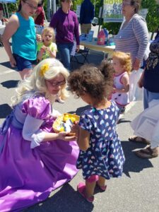 A woman dressed as a fairy tale character with long blond hair and a lilac gown is kneeling and handing out treats from an orange bucket to a young girl in a blue dress with heart patterns. Several other children and adults are standing nearby under a sunny sky.