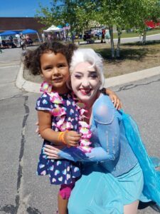 A young girl in a blue polka dot dress stands outside on a sunny day, smiling and wearing a colorful flower lei. She is hugging a woman dressed in a blue Elsa costume from the movie "Frozen" who is also smiling and kneeling beside her. Trees and tents are in the background.
