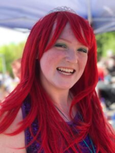 A person with red hair, wearing a small tiara and a sequined purple top, is smiling brightly. The background appears to be a sunny outdoor setting with blurred trees and a blue canopy visible.