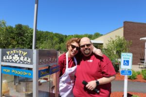 Two people are standing outdoors near a popcorn machine. The person on the left, wearing a white apron with a popcorn design, has an arm around the person on the right who is wearing sunglasses and holding a microphone. Both are smiling.