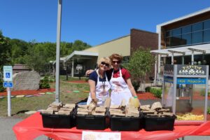Two people smile and pose behind a table with numerous popcorn bags in front of them. They are wearing aprons and sunglasses. A large popcorn machine is to their left. The setting appears to be outdoors in front of a building on a bright, sunny day.