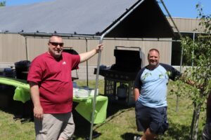 Two men stand outdoors under a canopy, next to a portable grill and a table covered with a green tablecloth. The man on the left wears sunglasses and a red shirt, while the man on the right wears a two-tone shirt. A building and part of another tent are in the background.