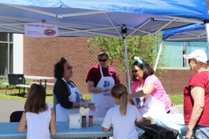 A group of women wearing aprons serve treats to two young girls under a blue canopy. One woman holds a container of toppings, while another scoops treats. A sign in the background reads "Allergic to nuts? No problem. We do that too!" A brick building is visible nearby.