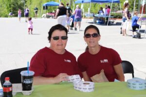 Two women wearing red t-shirts with white text sit at a table during an outdoor event. They are holding ticket rolls and smiling at the camera. A large drink cup and a soda bottle are on the table. People and booths are visible in the background.