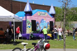 A colorful bouncy castle with a fairy tale princess theme stands in an outdoor area. People are gathered around, some under a white tent nearby. A bicycle is parked in the foreground, and a tree without leaves is to the right. The scene is sunny and vibrant.