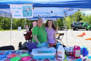 Two people standing under a canopy with a sign that reads, "Let's play with Bubbles!" They are in front of a table with various bubble-making supplies. The setting appears to be an outdoor event, with a van and trees visible in the background.