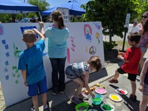 Children are painting on a large white board outdoors, using colorful paints. Some are using brushes and others are stamping with foam shapes. Adults stand nearby under blue tents, and a tree provides shade. The day is sunny, creating a cheerful atmosphere.