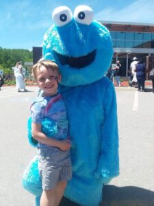 A smiling child in a tie-dye shirt and shorts stands outdoors next to a person in a blue furry costume with large eyes and a wide, open mouth, similar to Cookie Monster. Other people and a building are visible in the background.