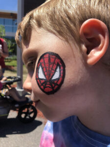 A close-up side view of a young child with a painted Spiderman face on their cheek. The child is looking down, and the background shows a building, sunlight, and a stroller with another child in it. The child is wearing a tie-dye shirt.