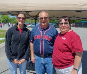 Three people smiling under a canopy on a sunny day. The individual in the center is wearing a Red Sox jersey, while the person on the left is dressed in a black jacket, and the person on the right is wearing a maroon shirt. All are standing on a paved area.