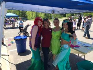 Four people are standing and smiling under a blue canopy at an outdoor event. Two are dressed as mermaids, one in a green leafy outfit, and another in a blue costume with a headband. A crowd and other booths are visible in the sunny background.