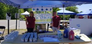 Two people stand under a blue canopy with a sign that reads, "Working Together for Strong Communities." The table in front of them holds cups, bottled water, pamphlets, and large jugs of liquid. Trees and other canopies are visible in the background.